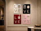 Four square wall art panels arranged in a grid, each featuring the word LOVE cut out in a bold, block-letter style. The panels are finished in a glossy glaze, with colors including white, black, deep red, and soft pastel pink.