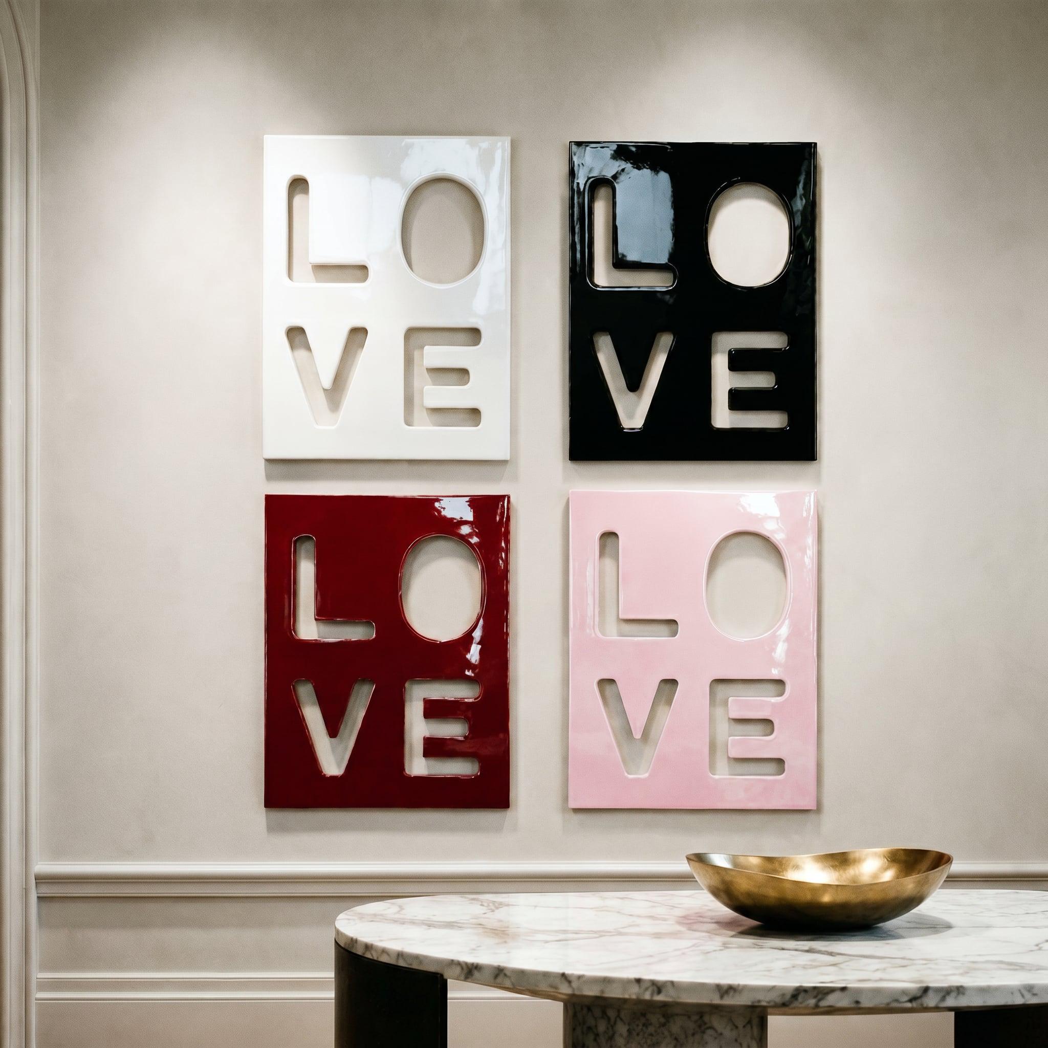 Four square wall art panels arranged in a grid, each featuring the word LOVE as a cutout. The top left panel is white, the top right is black, the bottom left is deep red, and the bottom right is light pink, all with a glossy finish.