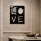 A square black wall plaque featuring the word LOVE cut out in a block letter design. The surface of the plaque is glossy and reflective, displaying the letters L, O, V, and E arranged in a two-by-two grid format on the wall.
