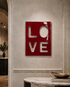 A rectangular wall art piece with a glossy, deep red finish featuring the word LOVE cut out in a block-letter style. The letters L, O, V, and E are arranged in a two-by-two grid, revealing the light-colored wall surface behind them.