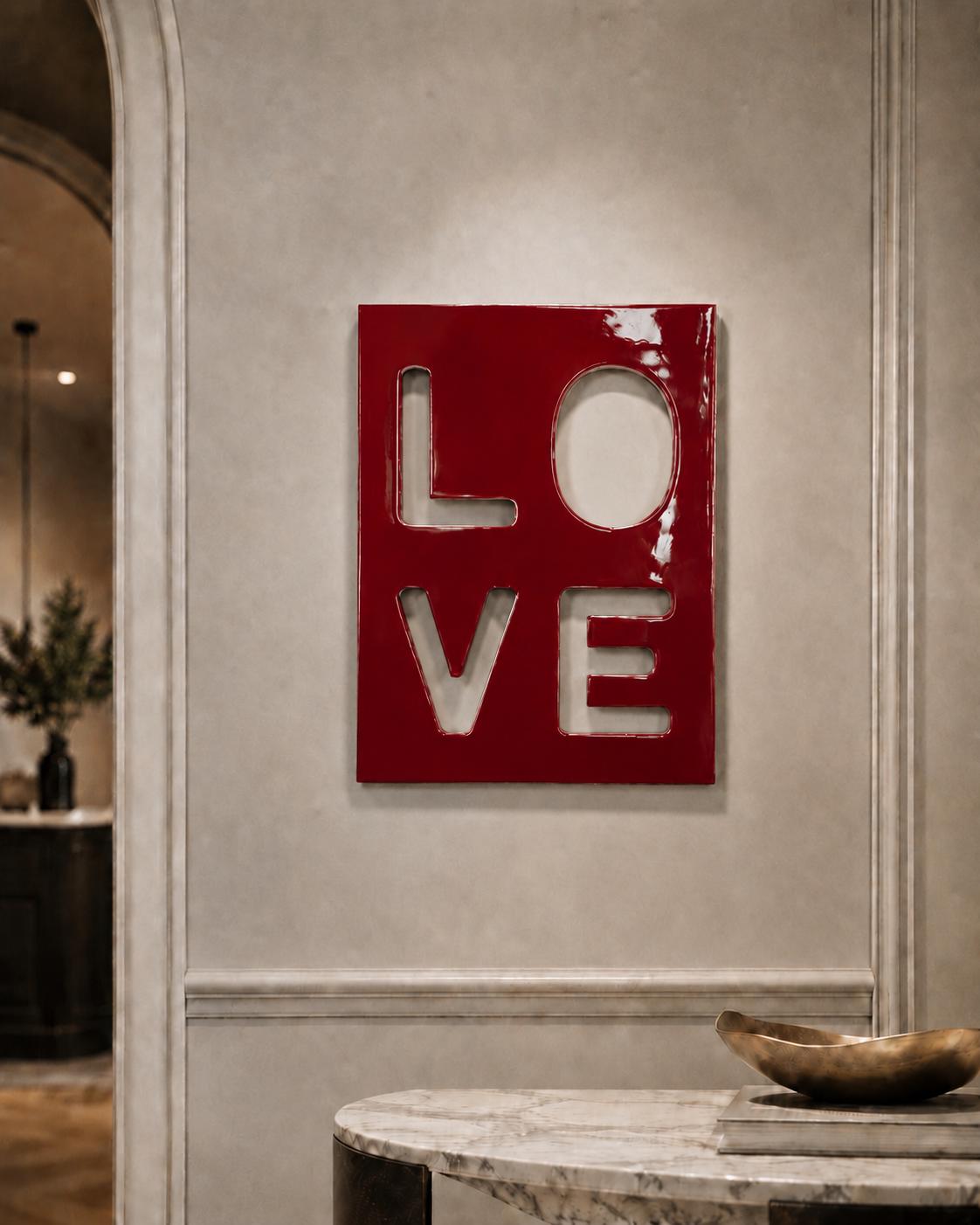 A rectangular wall art piece with a glossy, deep red finish featuring the word LOVE cut out in a block-letter style. The letters L, O, V, and E are arranged in a two-by-two grid, revealing the light-colored wall surface behind them.