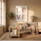 A large rectangular white wall art piece featuring the word LOVE in bold, recessed cut-out lettering. The artwork is displayed above a beige sofa with a chunky knit throw blanket, a wooden coffee table, and a potted olive tree.