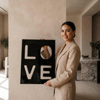 A rectangular black wall art piece featuring the word LOVE in large, cut-out block letters. The letters L, O, V, and E are arranged in a two-by-two grid, with the interior of each letter revealing a reflective mirror surface.