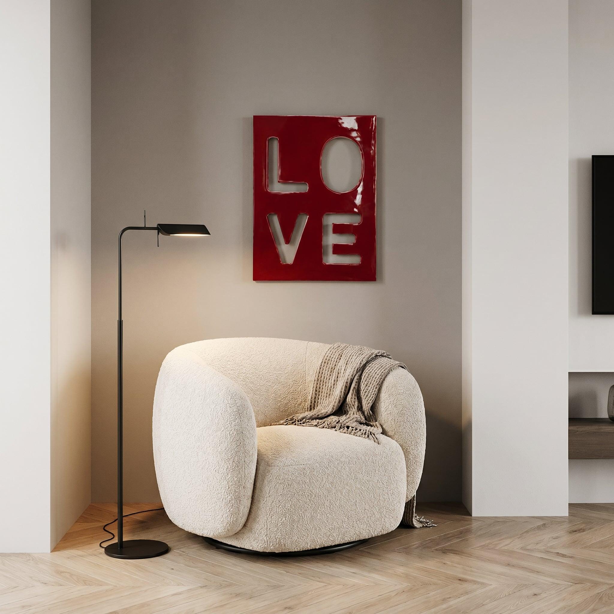 A cream-colored, rounded armchair with a textured fabric finish, draped with a beige throw blanket. Beside it stands a slim black floor lamp with a rectangular head. On the wall hangs a glossy red rectangular art piece with the word LOVE.