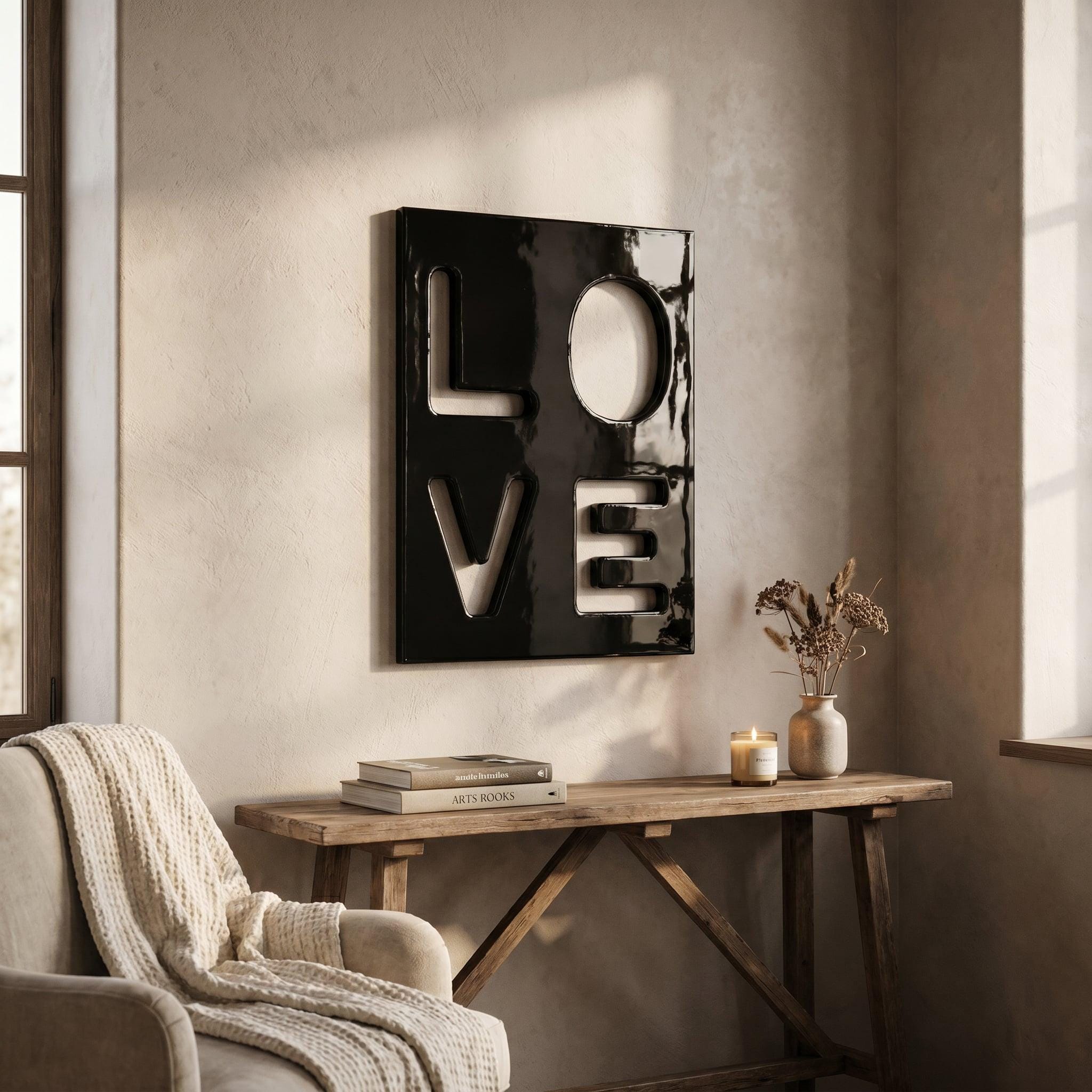 A square black wall art piece featuring the word LOVE cut out in a bold, block-letter style. The surface of the art is glossy and reflective, creating a high-contrast look against the wall. The letters L, O, V, and E are arranged in a two-by-two grid format, with the O appearing as a circular cutout.