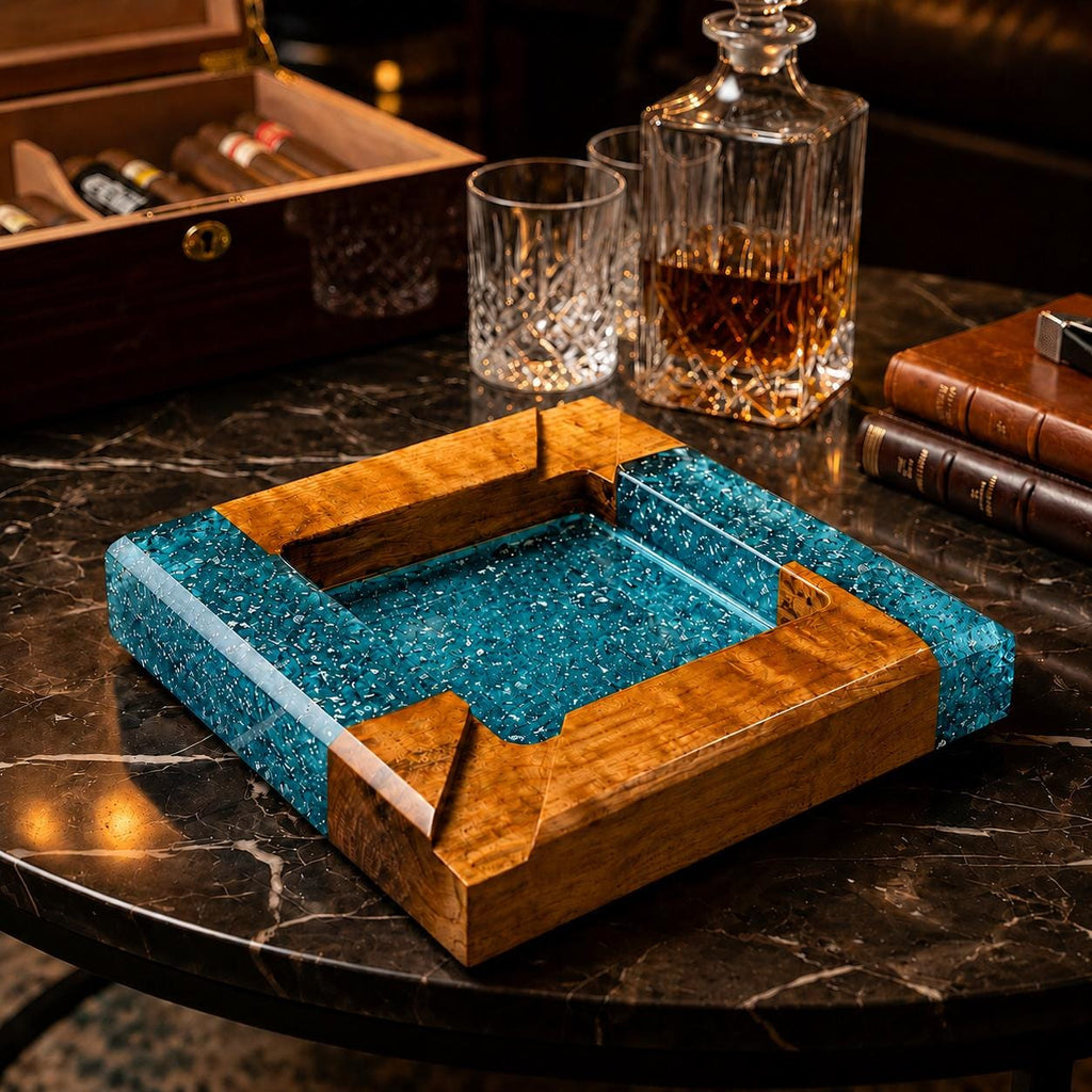 A square ashtray constructed from natural wood and translucent blue resin with embedded glitter particles. The design features a central square basin surrounded by a thick wooden frame with four notched corners for resting cigars.