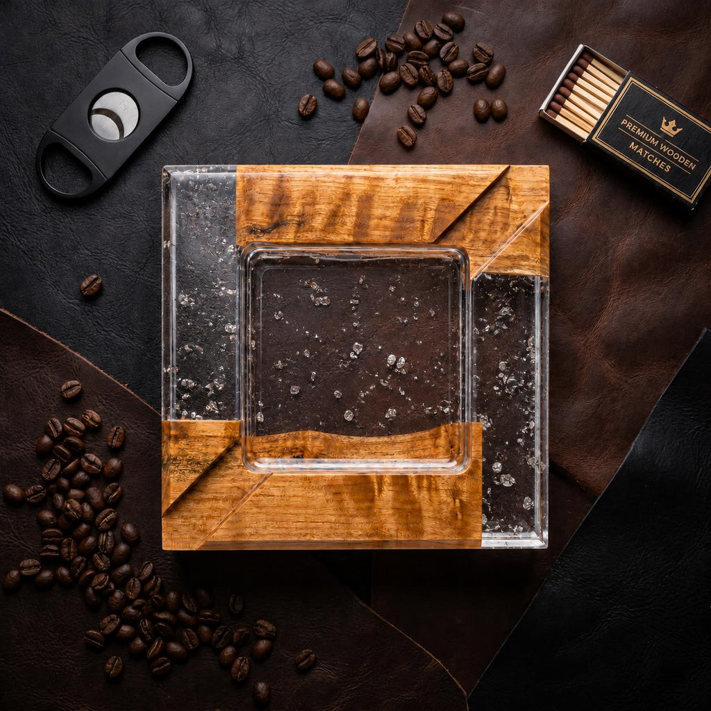 A square ashtray featuring a combination of natural wood and clear resin with embedded silver-colored flakes. The design includes a central square basin and four corner sections of wood. A black cigar cutter and a box of matches are nearby.