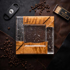 A square ashtray featuring a combination of natural wood and clear resin with embedded silver-colored flakes. The design includes a central square basin and four corner sections of wood. A black cigar cutter and a box of matches are nearby.