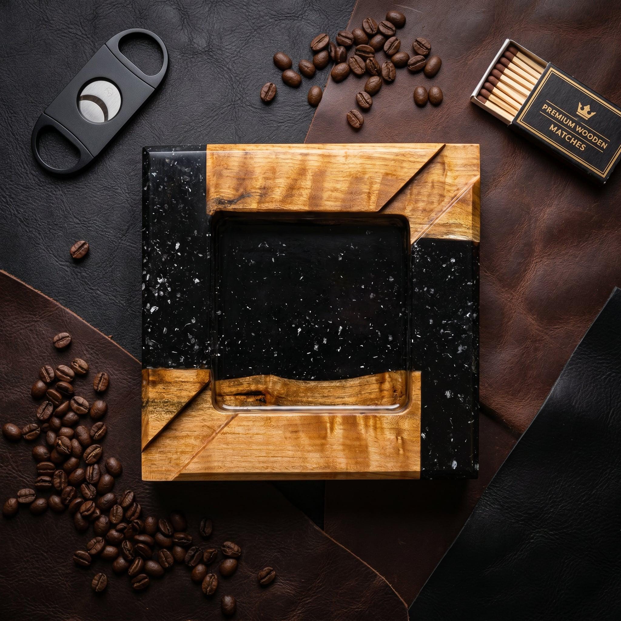 A square ashtray crafted from natural light-toned wood and black resin with white flecks. The design features a central square basin surrounded by a frame of alternating wood and resin segments. A black cigar cutter and a box of matches are nearby.