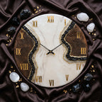 A round wall clock featuring a white and brown marbled face with a central jagged fissure filled with black crystals and gold accents. The clock has gold Roman numerals and thin black hands, surrounded by scattered black and white stones.