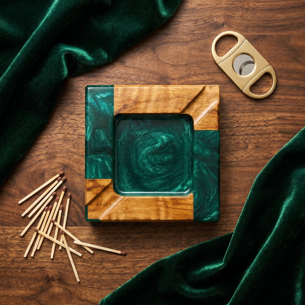 A square ashtray crafted from natural wood and swirling emerald green resin, featuring a central basin and two integrated cigar rests. A gold-toned metal cigar cutter and a small pile of wooden matches are positioned nearby.