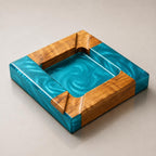 A square ashtray constructed from natural wood and vibrant turquoise resin. The design features a central recessed basin with a swirling pearlescent finish, framed by four wooden corner segments that include integrated notches.