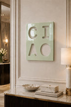 A square, light green wall plaque featuring the cutout letters C, I, A, and O arranged in a two-by-two grid. The surface of the plaque has a glossy, reflective finish, and the letters are formed by negative space against the wall.