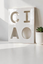 A square white ceramic plaque featuring the word CIAO cut out in a bold, block-letter style. The surface has a glossy finish, and the letters are arranged in a two-by-two grid. The plaque is positioned next to a small succulent plant.