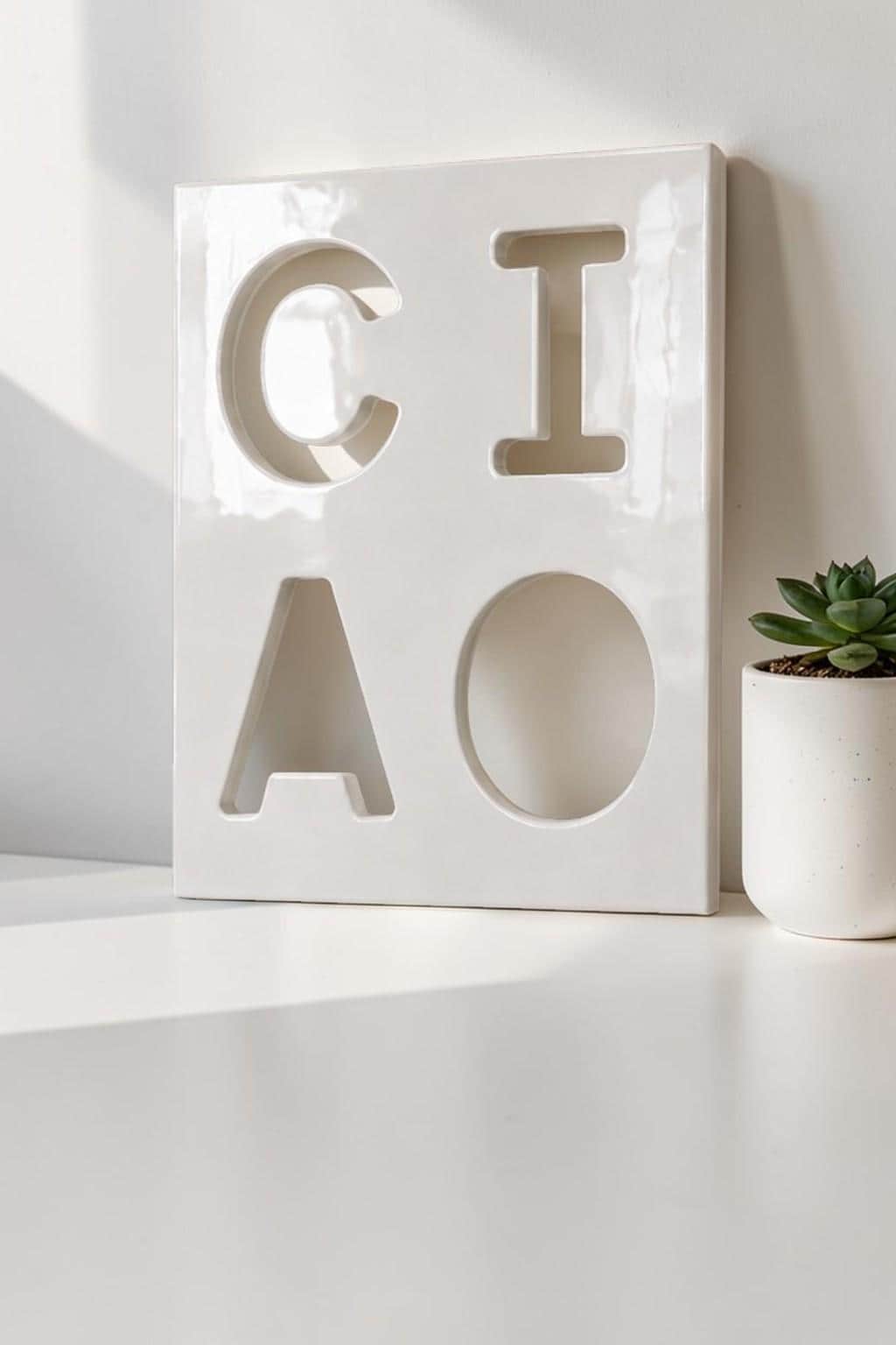A square white ceramic plaque featuring the word CIAO cut out in a bold, block-letter style. The surface has a glossy finish, and the letters are arranged in a two-by-two grid. The plaque is positioned next to a small succulent plant.