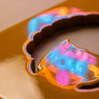 A smooth, glossy, mustard-yellow surface featuring a large, irregular circular cutout. The edges of the cutout reflect vibrant, iridescent light in shades of neon pink and electric blue, creating a high-contrast, reflective effect.