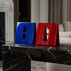 Two glossy, V-shaped decorative objects, one in vibrant blue and one in bright red. Each piece features a central vertical cutout with a horizontal bar, creating a pill-shaped opening. The objects have a smooth, reflective finish.