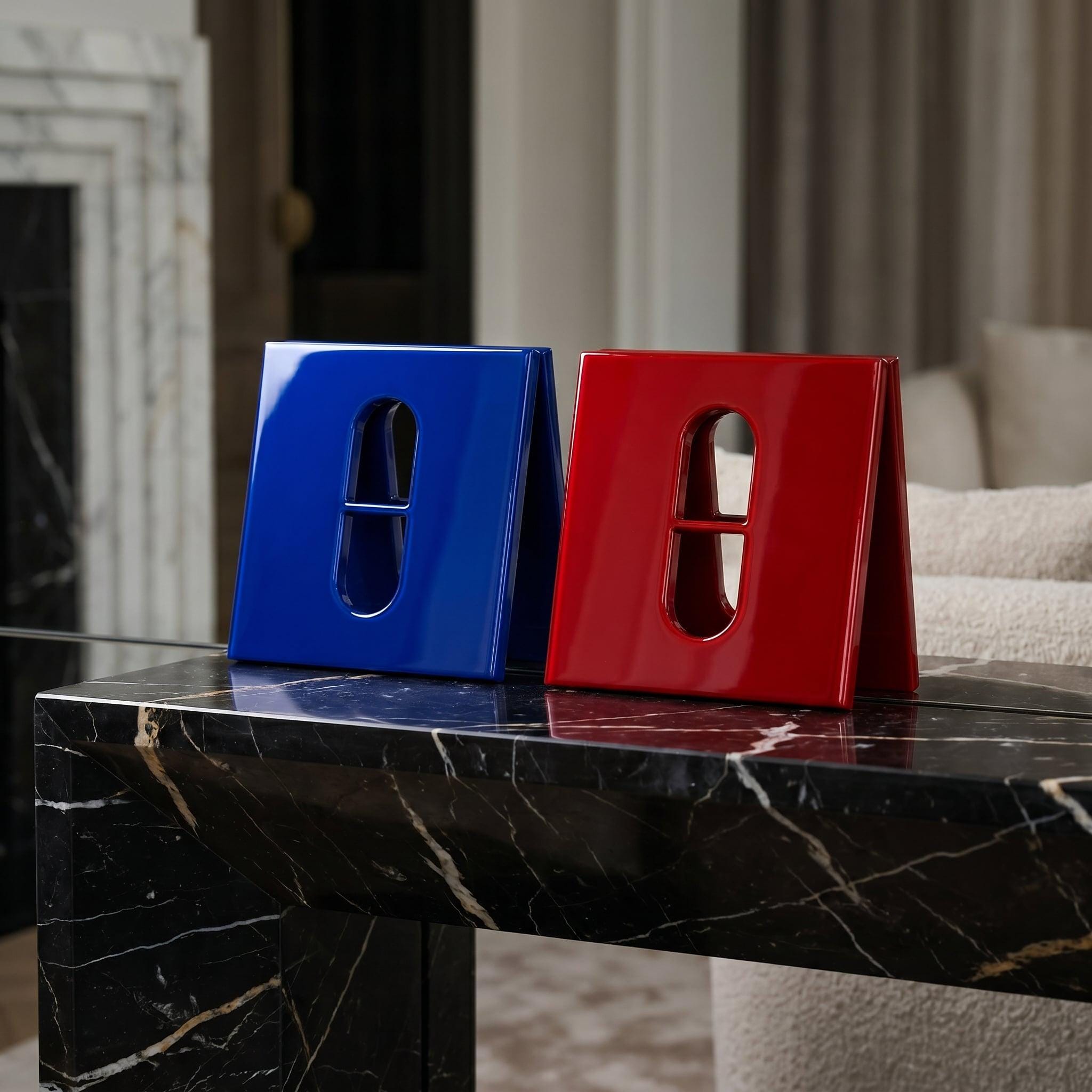 Two glossy, V-shaped decorative objects, one in vibrant blue and one in bright red. Each piece features a central vertical cutout with a horizontal bar, creating a pill-shaped opening. The objects have a smooth, reflective finish.