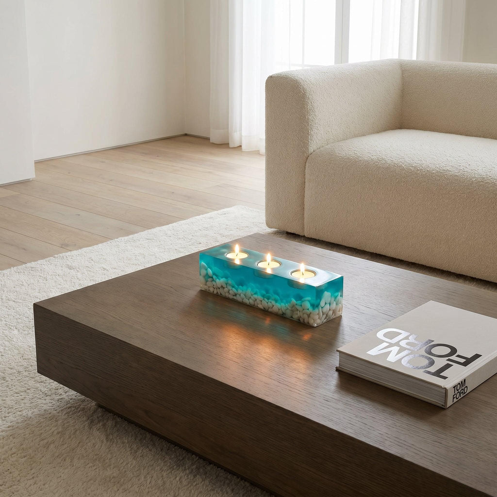 A rectangular candle holder made of clear resin with a vibrant turquoise blue tint, featuring embedded white pebbles at the base and three circular cutouts on top, each containing a small lit white tea light candle.