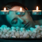 Three small round tea light candles with glowing orange flames are positioned in a horizontal row above a pile of smooth white river stones. The scene is illuminated by a cool teal light, casting soft shadows across the composition.