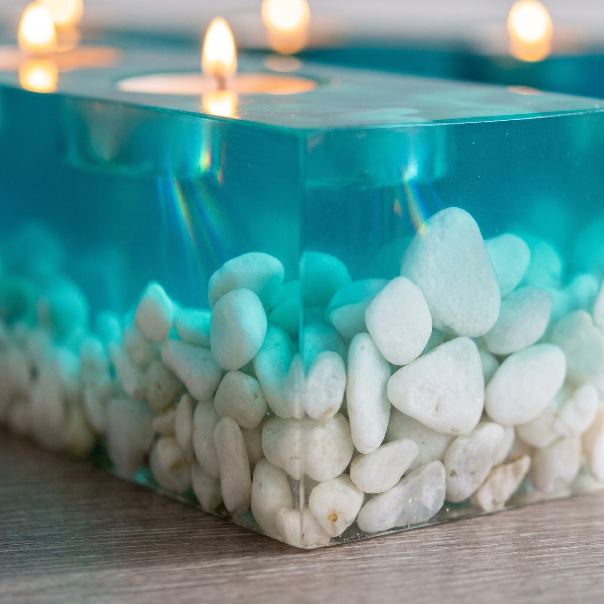 A rectangular teal resin candle holder featuring a clear, translucent top section and a base filled with numerous smooth, white river stones. The holder contains a lit tea light candle, casting a soft glow through the resin material.