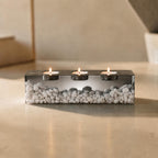 A rectangular clear resin candle holder containing numerous small white stones. The holder features three circular indentations along the top, each containing a lit tea light candle with a visible flame, creating a warm glow.