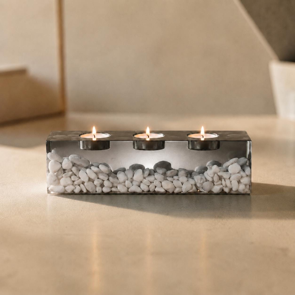 A rectangular clear resin candle holder containing numerous small white stones. The holder features three circular indentations along the top, each containing a lit tea light candle with a visible flame, creating a warm glow.