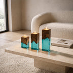 Three square candle holders of varying heights, each featuring a natural dark brown wood top section and a translucent turquoise blue resin base. Each holder contains a lit tea light candle centered in the top wooden surface.