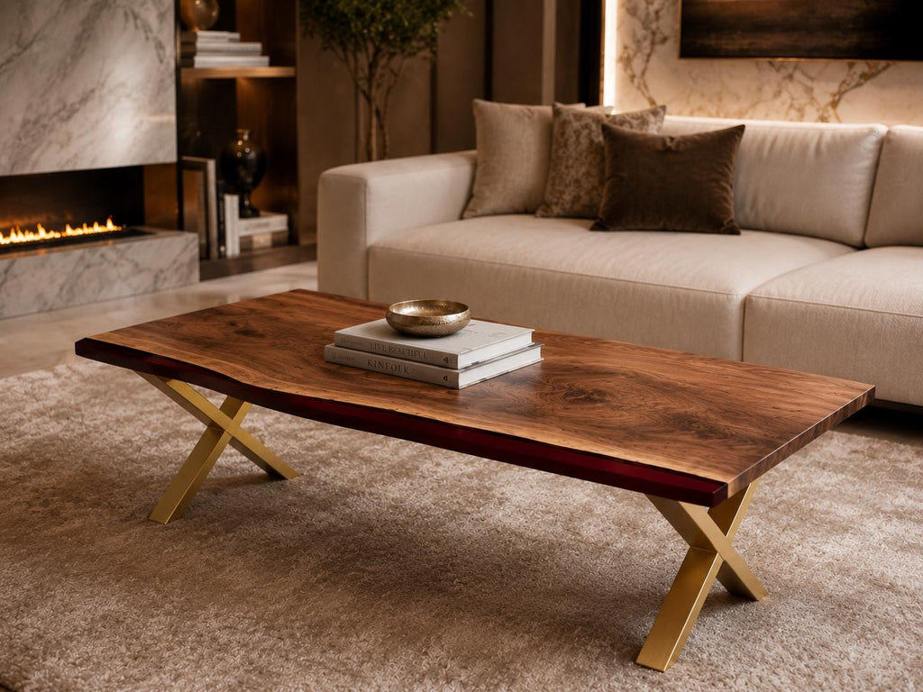 A rectangular coffee table featuring a natural live-edge dark wood slab top with a deep red resin inlay along one side. The table is supported by a gold-colored metal base with crossed legs, and it holds two stacked books and a small bowl.