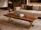 A rectangular coffee table featuring a natural live-edge dark wood slab top with a deep red resin inlay along one side. The table is supported by a gold-colored metal base with crossed legs, and it holds two stacked books and a small bowl.