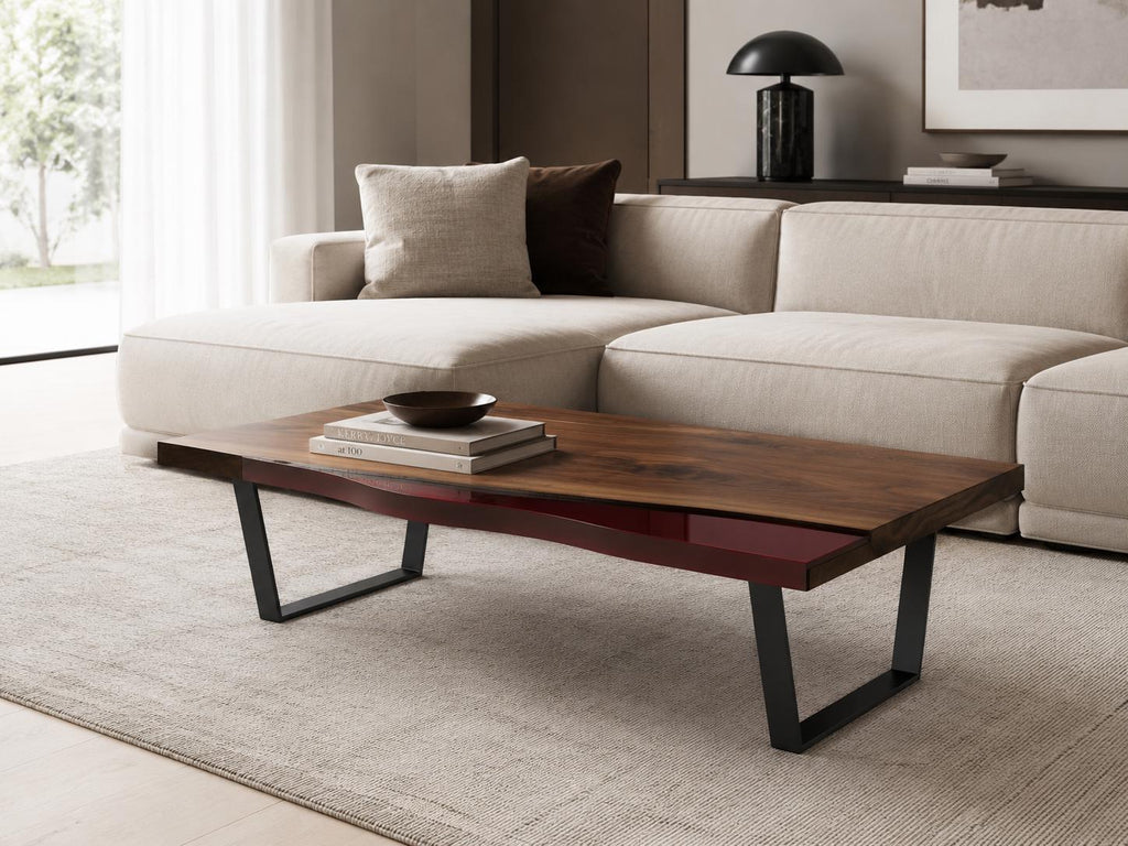 A rectangular coffee table featuring a natural wood grain top with a live edge, accented by a deep red resin inlay. The table is supported by two black metal U-shaped legs. A small dark bowl and two stacked books rest on the surface.