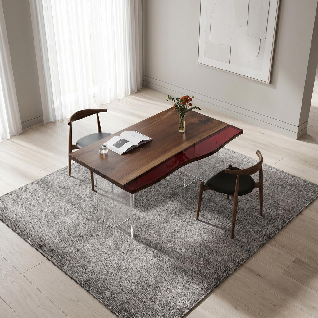 A rectangular dining table with a dark brown wood top featuring a wavy red resin inlay, supported by clear acrylic legs. Two wooden chairs with black upholstered seats are positioned at the table, which holds an open book.