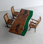 A rectangular dining table featuring a natural live-edge walnut wood slab with dark resin inlays and a solid deep green resin side section, supported by clear acrylic legs, accompanied by three wooden spindle-back chairs.