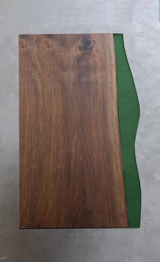 A rectangular table top featuring a dark brown wood grain surface on the left side and a smooth, solid green resin section on the right side. The green resin edge has a curved, organic shape that contrasts with the straight wood edge.