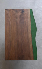 A rectangular table top featuring a dark brown wood grain surface on the left side and a smooth, solid green resin section on the right side. The green resin edge has a curved, organic shape that contrasts with the straight wood edge.
