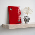 A glossy red rectangular plaque featuring two cut-out club symbols, one in the top left and one in the bottom right corner. Beside it sits a metallic silver geometric vase containing a single dried white palm leaf fan.