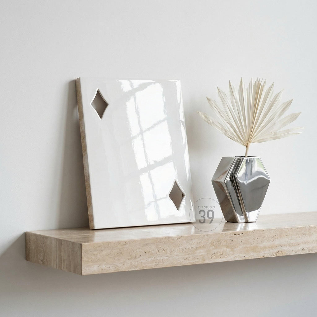 A glossy white rectangular plaque featuring two diamond-shaped cutouts, positioned next to a metallic silver geometric vase containing a dried palm leaf. The items rest on a beige travertine shelf with a natural porous texture.