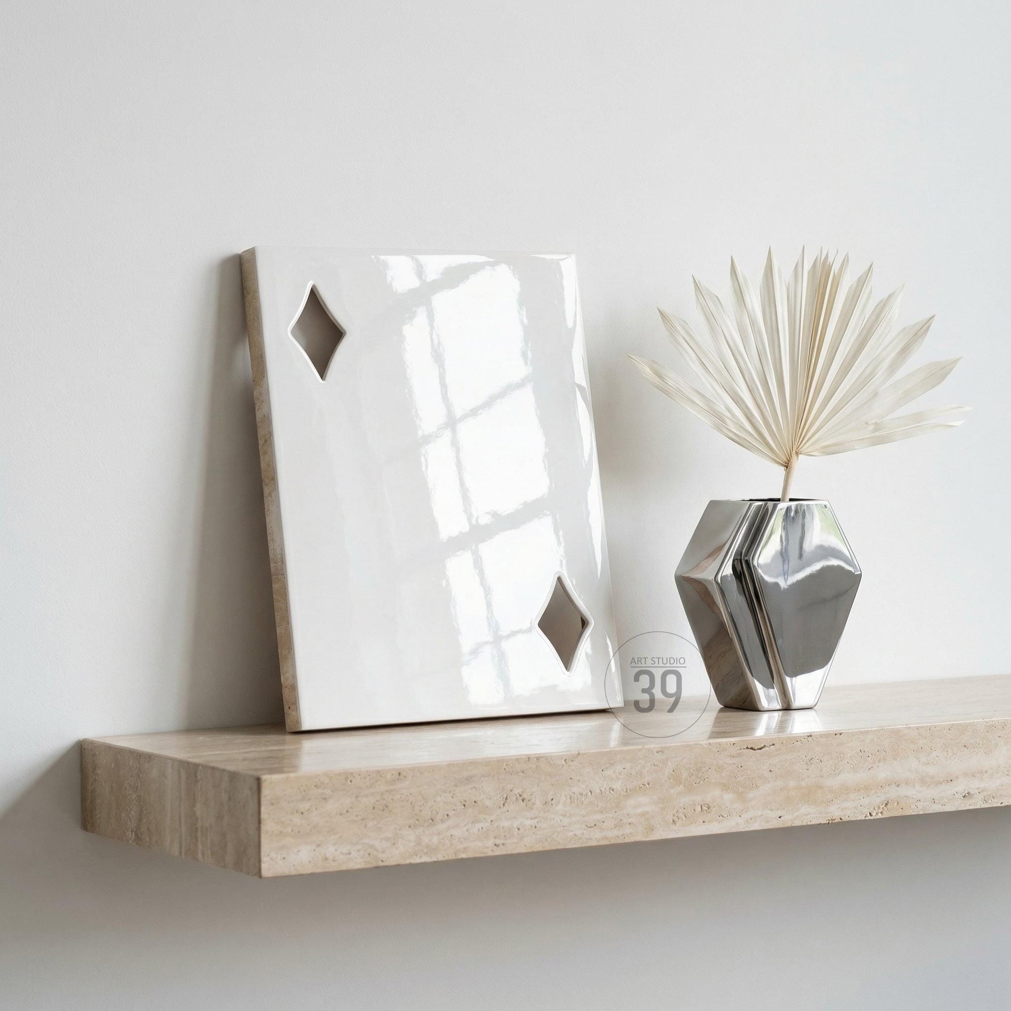 A glossy white rectangular plaque featuring two diamond-shaped cutouts, positioned next to a metallic silver geometric vase containing a dried palm leaf. The items rest on a beige travertine shelf with a natural porous texture.