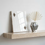A glossy white rectangular plaque featuring two diamond-shaped cutouts, positioned next to a metallic silver geometric vase containing a dried palm leaf. The items rest on a beige travertine shelf with a natural porous texture.