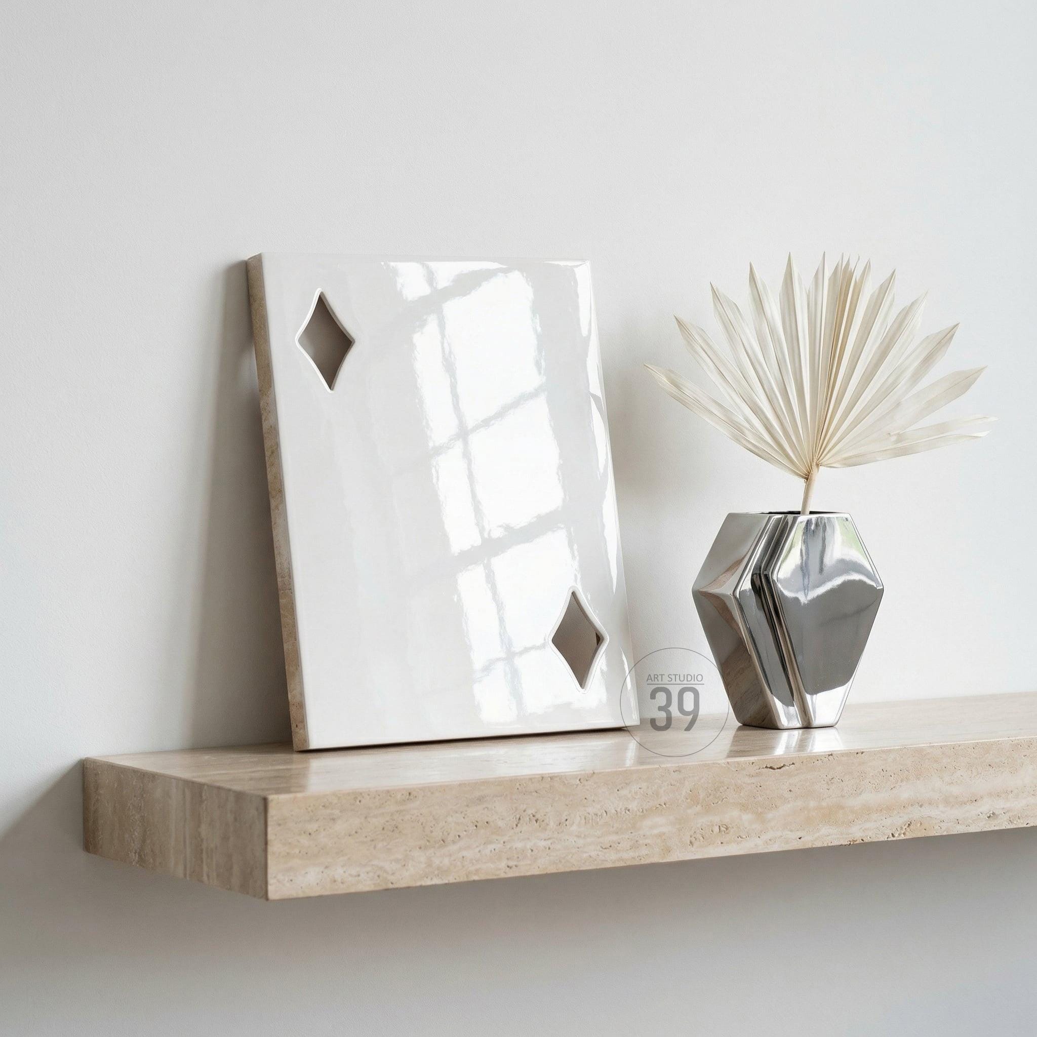 A glossy white rectangular plaque featuring two diamond-shaped cutouts, positioned next to a metallic silver geometric vase containing a dried palm leaf. The items rest on a beige travertine shelf with a natural porous texture.