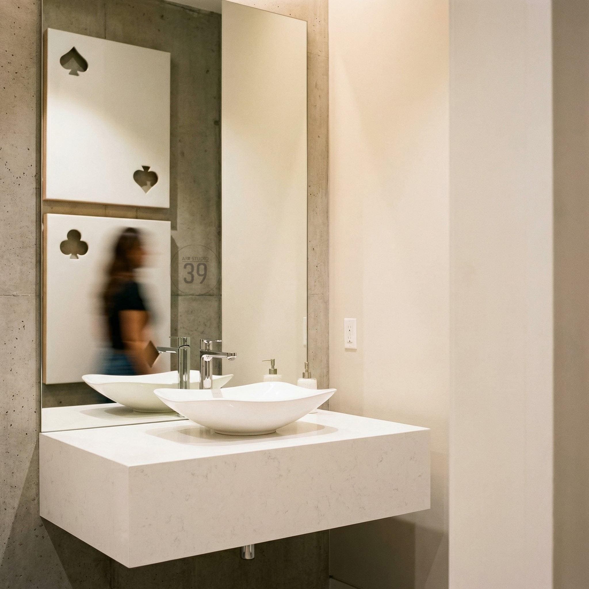 A white vessel sink sits on a floating white vanity countertop. A tall rectangular mirror above the sink reflects two white square wall panels, each featuring a cutout of a playing card suit, specifically a spade and a club.
