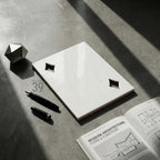 A rectangular white ceramic tile with two diamond-shaped cutouts, a black fountain pen, a black pen cap, a silver geometric polyhedral object, and an open book titled Modern Architecture: Form &amp; Function with architectural diagrams.