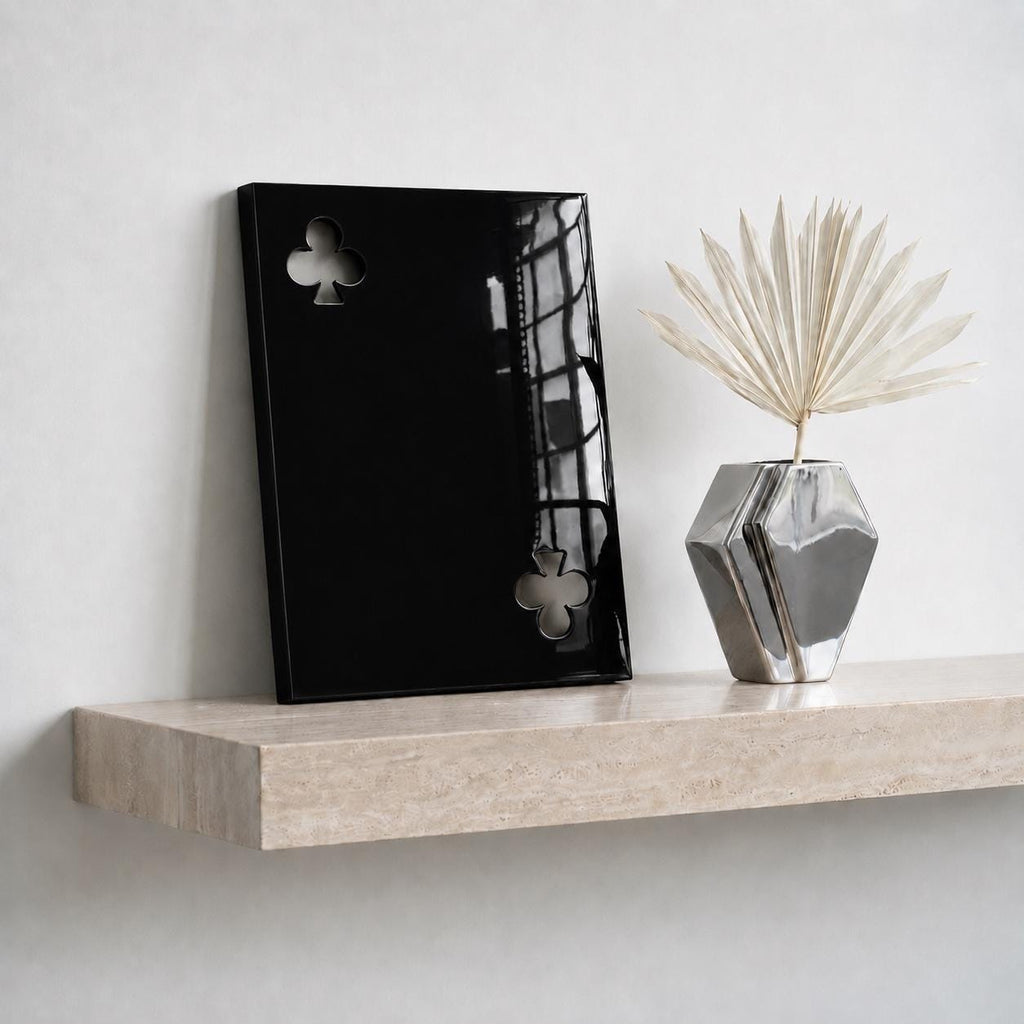 A rectangular black glossy plaque featuring two cut-out club symbols from a deck of cards, positioned next to a metallic silver geometric vase containing a single dried fan-shaped palm leaf on a light beige stone shelf.