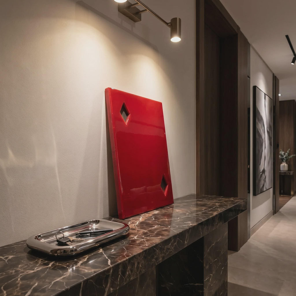 A rectangular red wall art piece featuring two diamond-shaped cutouts, positioned upright on a dark brown marble console table next to a polished silver metal tray containing a set of keys and a pen, illuminated by a wall-mounted light.
