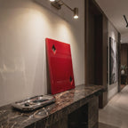 A rectangular red wall art piece featuring two diamond-shaped cutouts, positioned upright on a dark brown marble console table next to a polished silver metal tray containing a set of keys and a pen, illuminated by a wall-mounted light.