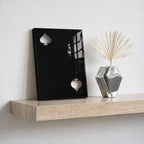 A rectangular black plaque featuring two cut-out spade symbols, one in the upper left and one in the lower right corner. Beside it sits a faceted silver vase containing a single dried, fan-shaped palm leaf in a light beige color.