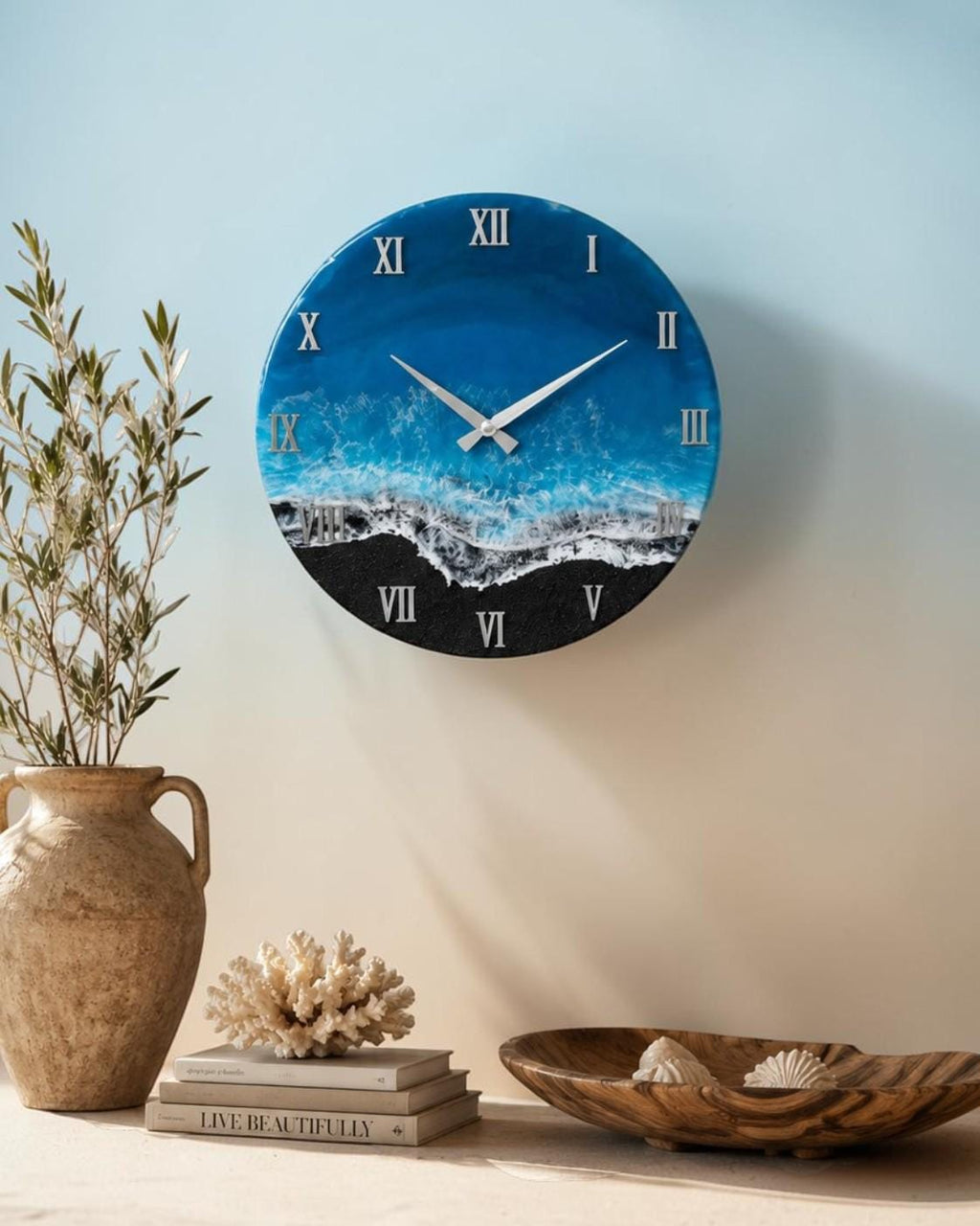 A round wall clock featuring a resin art design that mimics an ocean scene with deep blue water, white sea foam, and a black sand beach. The clock face displays silver Roman numerals and matching silver hour and minute hands.