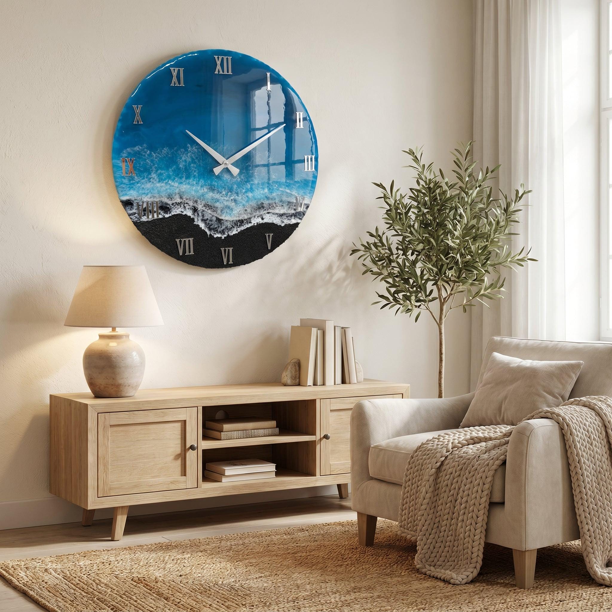 A circular wall clock featuring a glossy resin design that mimics an ocean shoreline, with deep blue water transitioning into white sea foam and a black sand beach. The clock face displays silver Roman numerals and thin silver hands.