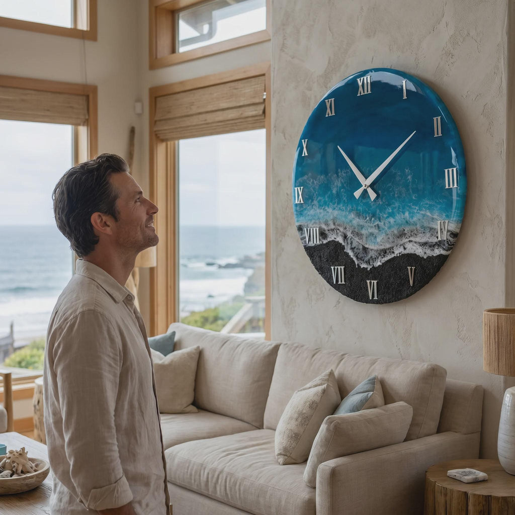 A large circular wall clock featuring a glossy resin finish that mimics an ocean scene with deep blue water, white sea foam, and a black sandy beach. The clock face displays silver Roman numerals and thin silver clock hands.