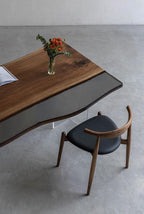 A wooden desk with a live edge and a gray glass insert, featuring a clear glass vase with orange flowers and a book on top. Beside the desk is a wooden chair with a black upholstered seat and a curved backrest.